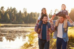 A young couple each carrying a child on their shoulders walking alongside a lake at sunset.